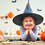 A little girl in a witch hat surrounded by Halloween treats and decorations.