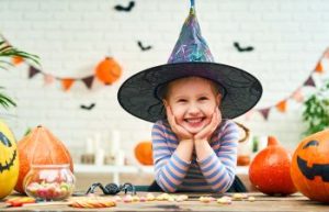 A little girl in a witch hat surrounded by Halloween treats and decorations.