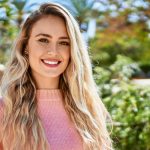 A woman with long blonde hair wearing a pink sweater stands outdoors, smiling at the camera with greenery and sunlight in the background.