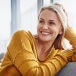 A woman in a yellow sweater sits on a couch, smiling and looking to her left with her hand resting on her head.