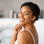 Natural Beauty. Attractive Black Woman Touching Chin And Looking At Camera While Standing In Bathroom At Home, Beautiful African American Female Enjoying Her Smooth Skin After Skincare Routine
