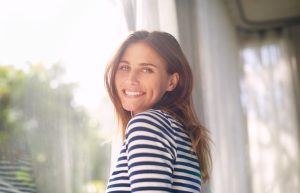 Woman, smile and portrait in house for relax and calm for holiday and cheerful on blurred background. Young person and happy with window for view of garden or backyard with sunlight in lounge