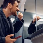 Happy man brushing his teeth while looking himself in am mirror.