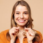 Young fun woman wear orange shirt casual clothes hold in hand invisible transparent aligners, invisalign bracer smile isolated on plain pastel light beige background studio portrait. Lifestyle concept