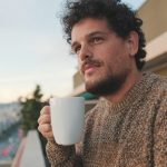 Peaceful guy looks at the city from the balcony and drinks coffee, enjoys the beautiful view