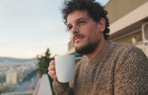 Peaceful guy looks at the city from the balcony and drinks coffee, enjoys the beautiful view